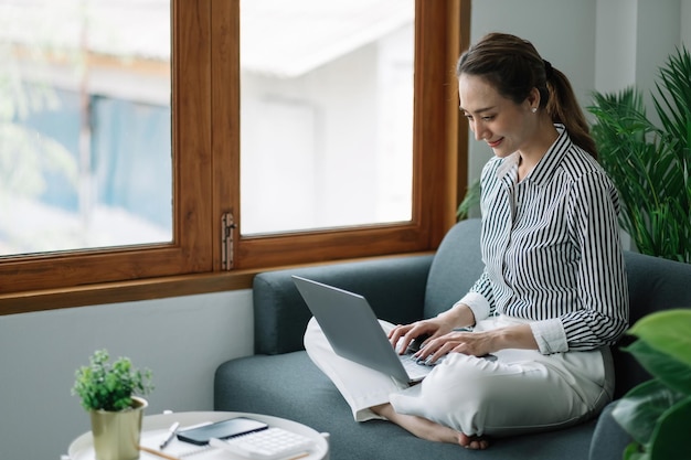 Азиатская молодая женщина, работающая из домашнего офиса, сидит на диване и держит бумажный счет-фактуру, проверяет информацию онлайн