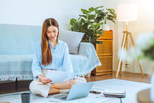 Giovane donna asiatica che lavora da casa usando il computer e bevendo caffè nella sua stanza documenta la finanza e la conferenza online per nuovi progetti