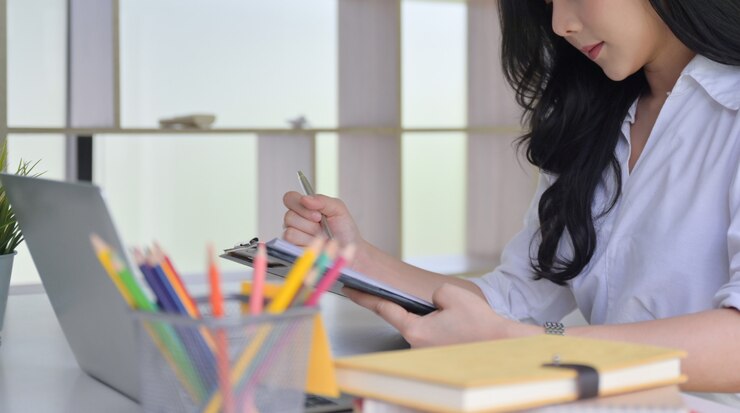 Photo 亞洲年輕女子在辦公室工作, 她看著手裡的文件, 桌上有筆記型電腦和辦公用品.