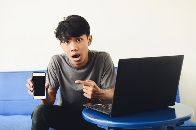 Asian youth sits on the sofa in front of the laptop pointing at his cell phone