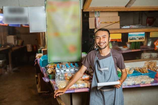 Asian youth wearing an apron stands holding a notebook at a traditional stall