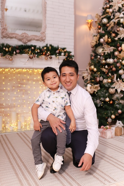 Foto papà e figlio asiatici ridono, sorridono e si abbracciano davanti al camino e all'albero di natale a casa