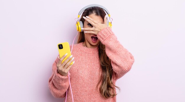 Asiatic pretty woman looking shocked, scared or terrified, covering face with hand. headphones and phone concept