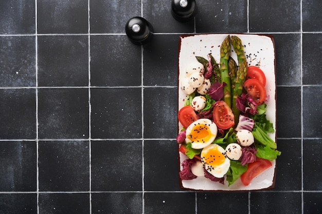 Asparagus tomato lettuce mozzarella black sesame flax oil olive salad and soft boiled egg on rectangular ceramic plate on black tile table background Healthy and diet food concept Top view