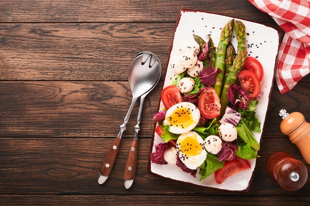 아스파라거스 토마토 상추 모짜렐라 검은 참깨 아마 기름 올리브 샐러드와 어두운 나무 테이블 배경에 직사각형 세라믹 접시에 부드러운 삶은 계란 건강 및 다이어트 식품 개념 상위 뷰