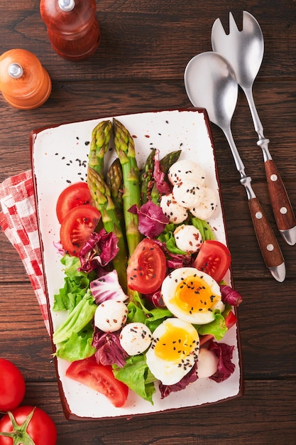 Asparagus tomato lettuce mozzarella black sesame flax oil olive salad and soft boiled egg on rectangular ceramic plate on a dark wooden table background Healthy and diet food concept Top view