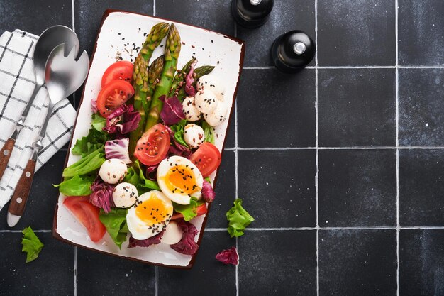 Asperges tomaat sla mozzarella zwarte sesam vlas olie olijf salade en zacht gekookt ei op rechthoekige keramische plaat op zwarte tegel tafel achtergrond gezond en dieet voedsel concept bovenaanzicht