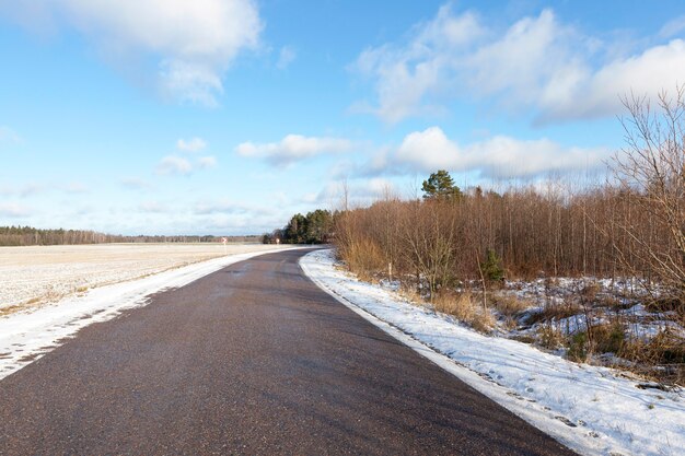 Asphalt road that separates the forest and agricultural field. Blue sky.