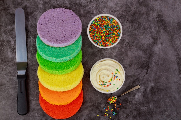 Photo assembling rainbow layer cake with white cream and sprinkles.