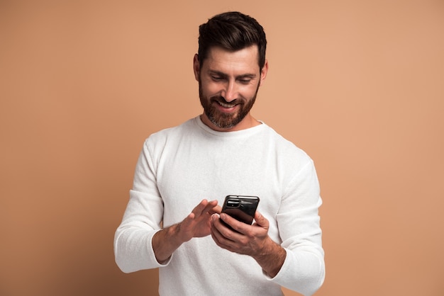 Assertive bearded man attentively looking at display of smartphone he holding, surfing internet, doing shopping online. Indoor studio shot isolated on beige background