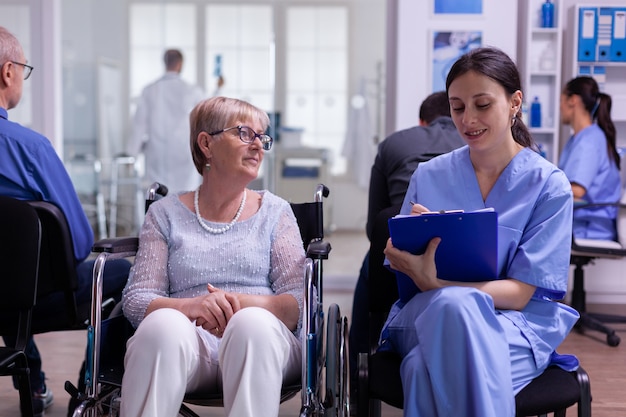 Assistente che prende appunti negli appunti sui problemi di salute del paziente in attesa di un medico specialista seduto su una sedia a rotelle nella sala d'attesa della clinica ospedaliera