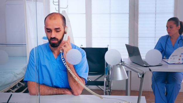 Assistant talking at phone, checking appointment sitting behind a glass wall in hospital office. Medical physician in medicine uniform writing list of consulted, diagnosed patients, making research.