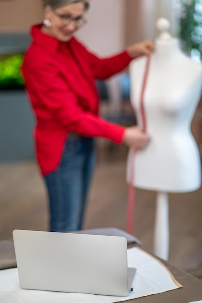 Foto assistente. computer portatile funzionante sul tavolo e donna in piedi manichino di misurazione guardando lo schermo in atelier