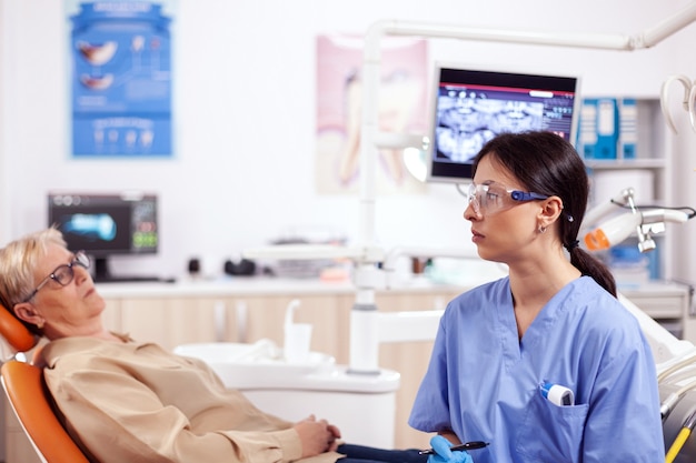 Assistent in tandartskabinet tijdens controle van de mondhygiëne van oudere vrouwen zittend op een stoel. senior vrouw in gesprek met medisch verpleegkundige in stomatologie kantoor over tanden probleem.