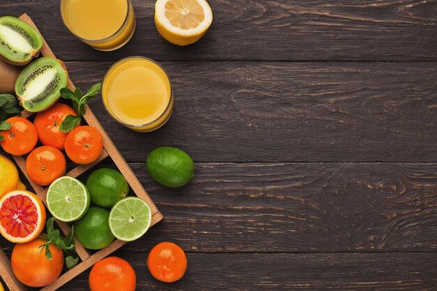 Assorted citrus fruits and fresh juice glasses for breakfast on dark wooden background. Top view on box with oranges, lemons, tangerines and other exotic fruits, flat lay, copy space