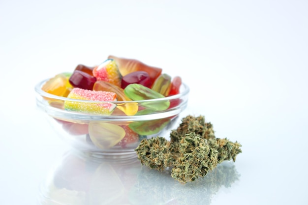 Assorted different gummy candies in a small glass bowl near in the foreground several buds of dried medical marijuana reflected on the glass