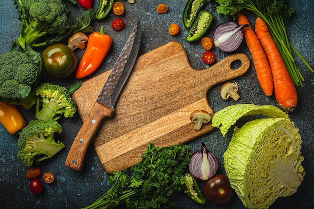 Assorted fresh vegetables on rustic concrete table with wooden cutting board in center and kitchen knife