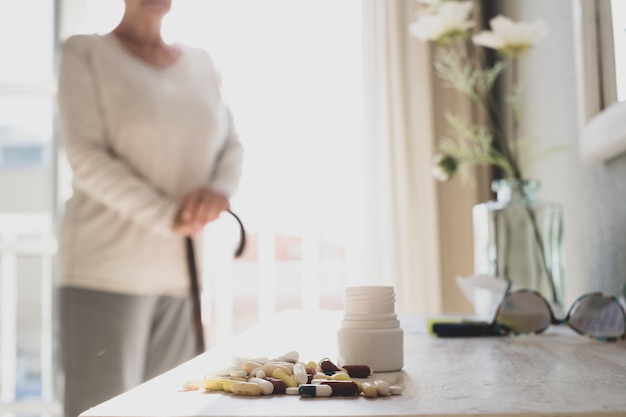 Assorted multicolored tablets, pills, capsules with bottle on table while senior woman standing in background at home. Old woman holding walking stick with pills and capsules on table at home