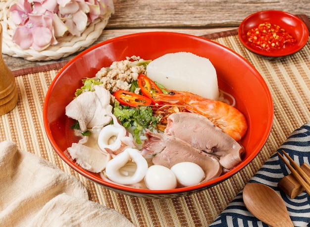 Assorted noodles soup with shrimp squad and vegetable in a bowl\
with spoon and chopsticks isolated on mat side view on wooden table\
taiwan food