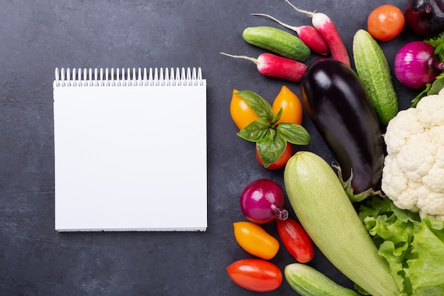 Assorted raw organic vegetables and notebook on dark stone background