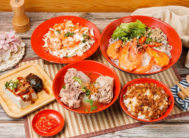 Assorted taiwan food salad with fresh shrimp braised pork rice\
stewed pork ribs noodles preserved egg tofu in a bowl with spoon\
and chopsticks isolated on mat side view on wooden table taiwan\
food