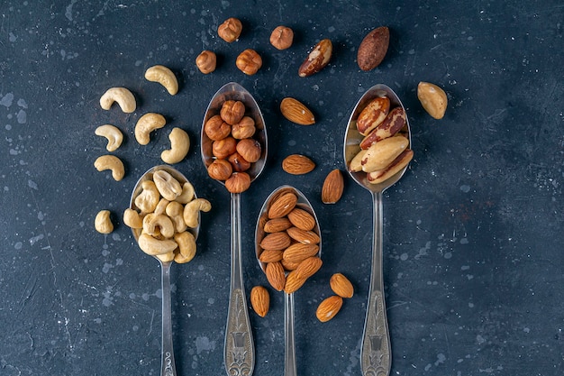 Assortiment van verschillende soorten noten: cashewnoten, hazelnoten, amandelen, paranoten op metalen zilveren lepels op donkere achtergrond