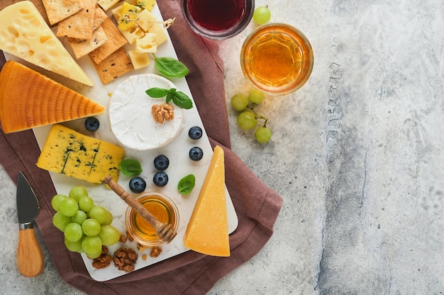 Assortment of cheese honey cracker blueberries grapes with red and white wine in glasses antipasto server on white marble board on grey background Flat lay copy space