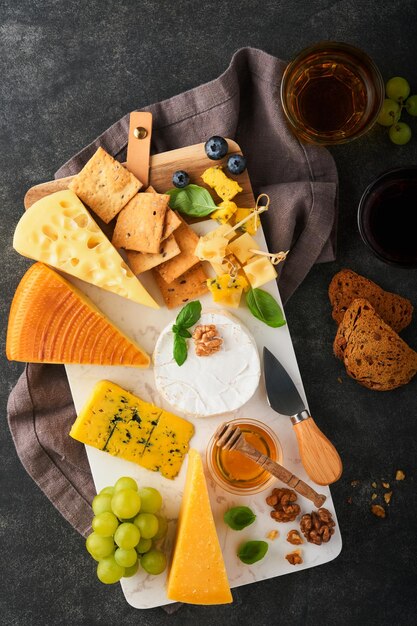 Assortment of cheese honey cracker blueberries grapes with red and white wine in glasses antipasto server on white marble board on old dark grey background Flat lay copy space