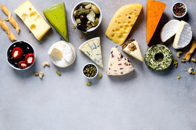 Assortment of hard, semi-soft and soft cheeses with olives, grissini bread sticks, capers, grape, on grey concrete backgound. Top view, copy space, flat lay. Cheese selection appetizer plate.