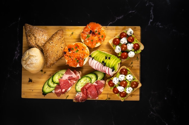 An assortment of sandwiches with fish, cheese, meat and vegetables lay on the board and a bun