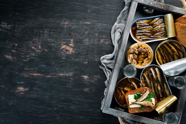 Assortment of sprats and snacks In a wooden box On a black background top view Free copy space