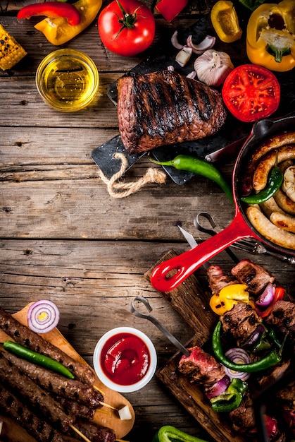 Assortment various barbecue food grill meat, bbq party fest - shish kebab, sausages, grilled meat fillet, fresh vegetables, sauces, spices, on old wooden rustic table, above copy space