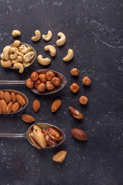 Assortment of various types of nuts: cashew, hazelnuts, almonds, brazil nuts on metal silver spoons on dark background