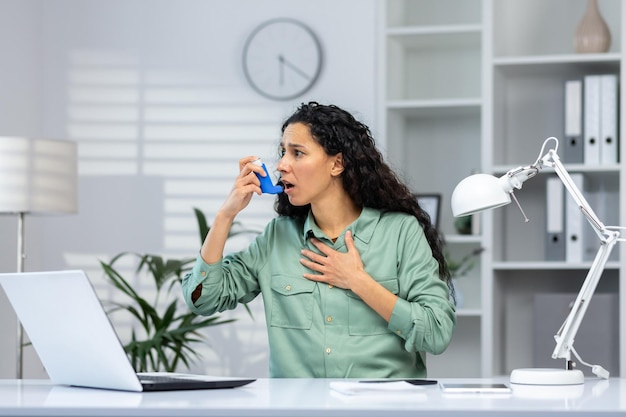 Astma-aanval bij Spaanse zakenvrouw binnen kantoor op werkplek met laptop vrouw gebruikt inhalator