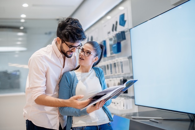 Astonished couple looking at specifications for smart TV they want to buy. They standing in front of TV.