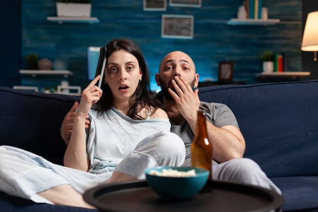 Astonished couple watching movie at night and eating popcorn drinking beer