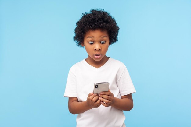 Astonished cute little boy with curly hair reading message on smartphone and expressing amazement shock, surprised by mobile application, using cellphone. studio shot isolated on blue background