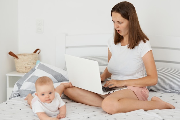 Photo astonished dark haired woman sitting on bed with her infant daughter and working online holding laptop and looking at display with open mouth expressing shock