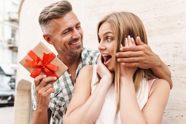 astonished excited couple in summer clothes smiling and holding present box together while standing against wall on city street