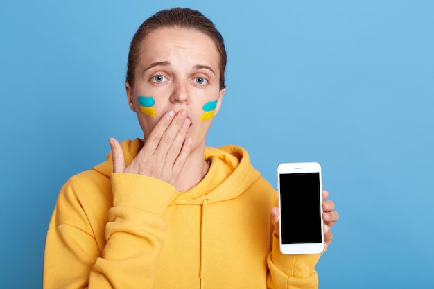 Photo astonished shocked woman in yellow hoodie with ukrainian flag on cheeks posing isolated over blue background covering mouth with palm showing cell phone with blank black screen