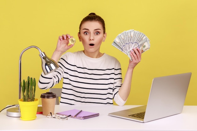 Astonished woman holding and showing fan of hundred dollar banknotes and bitcoin crypto coin