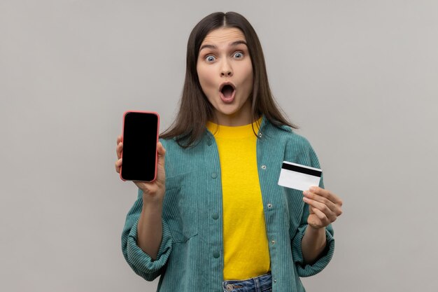 Astonished woman with dark hair holding credit card and smart phone with blank black screen