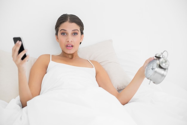 Astonished young brown haired model in white pajamas holding a mobile phone and an alarm clock