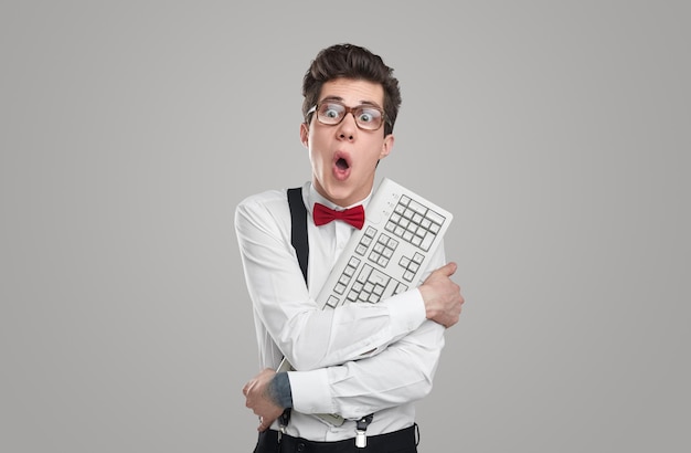 Astonished young man in glasses embracing computer keyboard and looking away while working on code against gray background