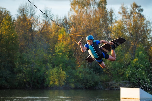 An athlete does a trick from a springboard A rider jumps on a wakeboard against a background of a green forest Sunset on the lake