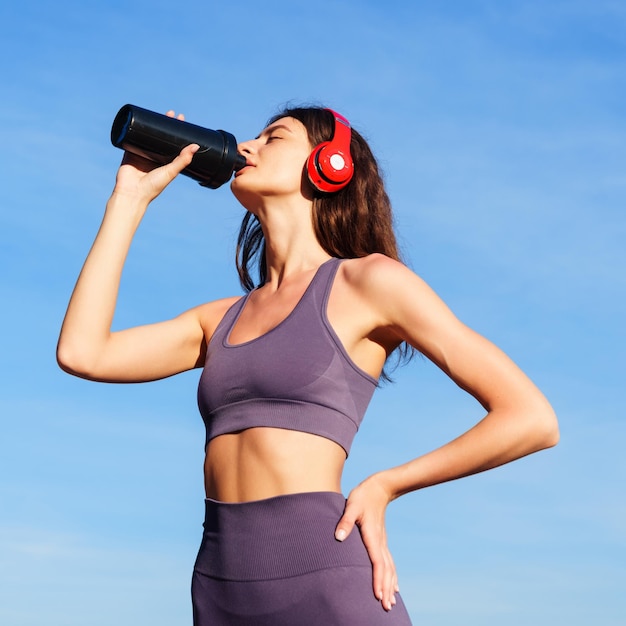 An athlete drinks water from a bottle against a blue sky Training outside in the morning