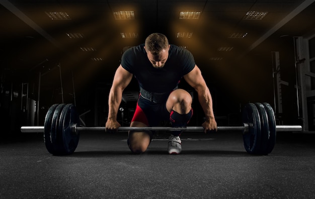 Athlete is standing on his knee and near the bar in the gym and is preparing to make a deadlift