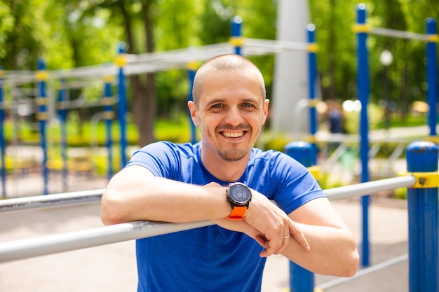 Athlete man leaning on bars at sports ground looking in camera and smiling after outdoor workout