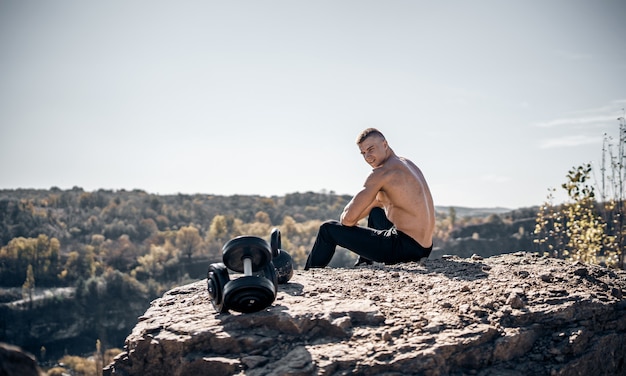 Athlete man on a rocky background. Perfect body. Quarry or mountain background. Sitting on a rock. Dumbbels on front view.
