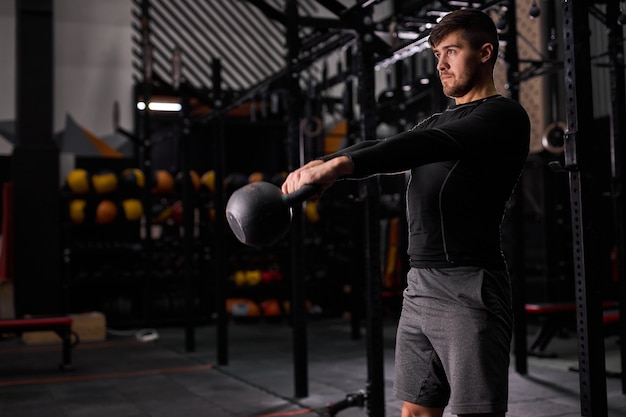 Athlete man in sportswear doing exercises with kettlebell while standing in cross fit gym, pumping arm muscles, training alone. sport, weightlifting concept